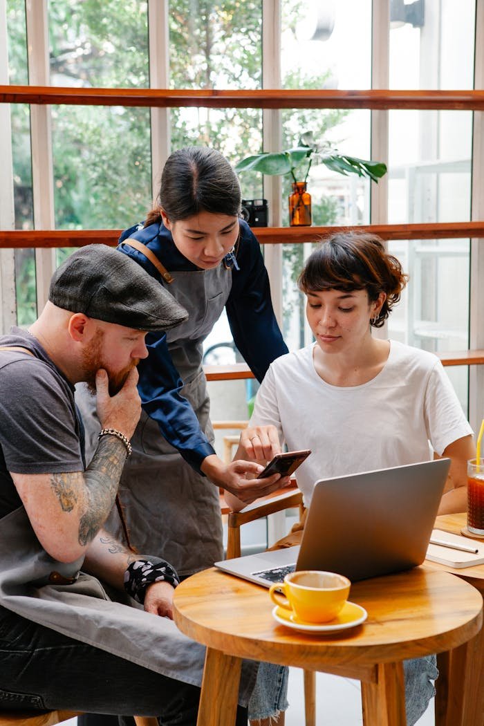 Group of coworkers in aprons and casual clothes using computer and cellphone at table in cafe with coffee cup in day